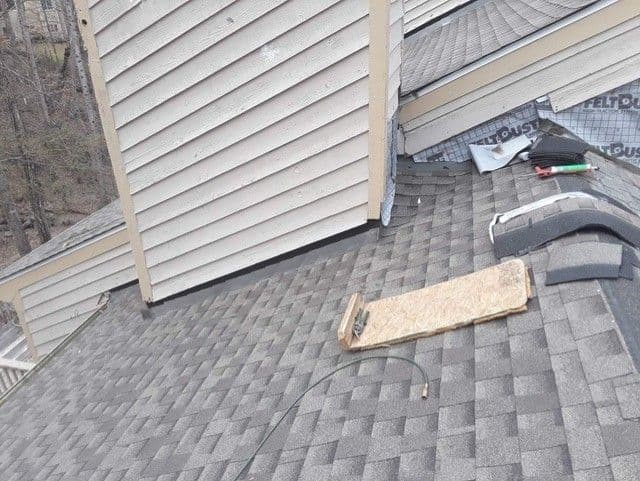 A shingled roof with a chimney and an adjacent wall. A piece of plywood, a coil of hose, and roofing materials are scattered on the surface. The scene suggests ongoing roof maintenance or repairs amidst overcast weather.