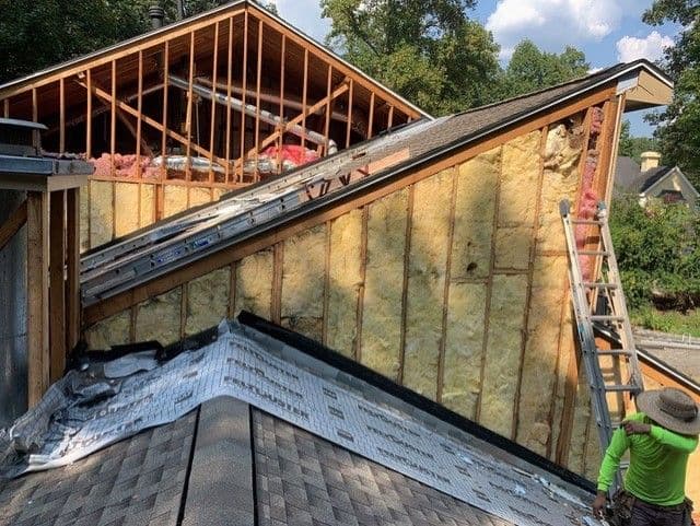 Alt text: "Roofing construction scene with exposed wooden beams and insulation. A worker in a green shirt stands near a ladder beside the house under a clear sky."