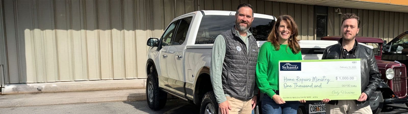 Three people stand smiling in front of a white truck, holding a large check for $1,000 from Shantz. The atmosphere is cheerful and bright.