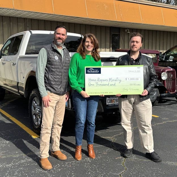 Three people stand smiling in front of a white truck, holding a large check for $1,000 from Shantz. The atmosphere is cheerful and bright.