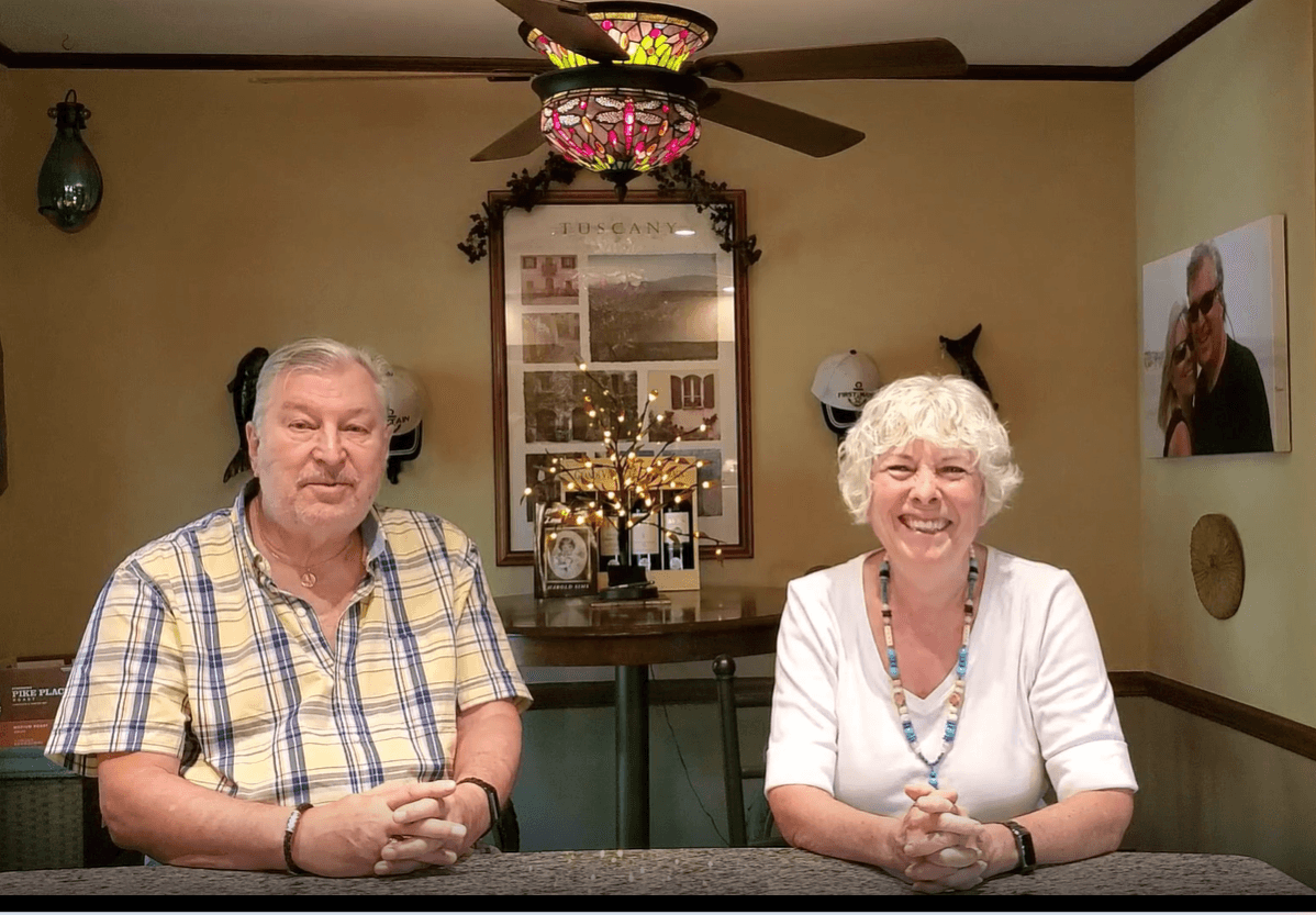 A man and woman sit at a table, smiling warmly. Behind them are decorative elements including a colorful light fixture, framed pictures, and small plants. The atmosphere is cozy and inviting.