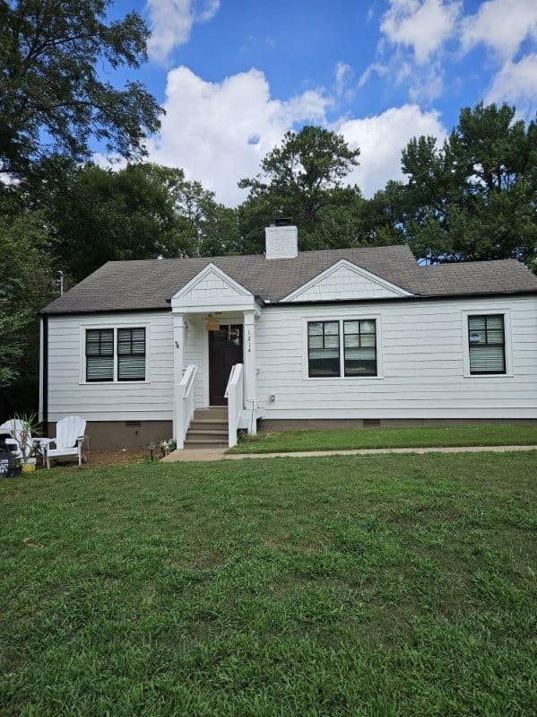A quaint, single-story white house with dark roof shingles sits amid lush green grass. Trees frame the scene, creating a serene and inviting atmosphere.