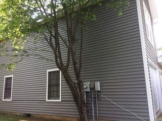 Side view of a grey house with two windows, featuring a tree in the foreground. Utility meters are mounted on the wall. The scene is calm and quiet.