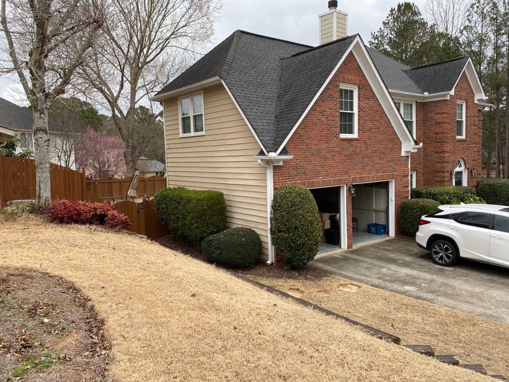 Suburban two-story house with red brick and beige siding, surrounded by bushes. A white car is parked in the driveway. Overcast sky creates a calm mood.