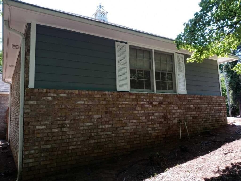 A house exterior with brick lower walls and dark gray-blue siding on top. Large windows with white shutters are framed by lush green foliage.
