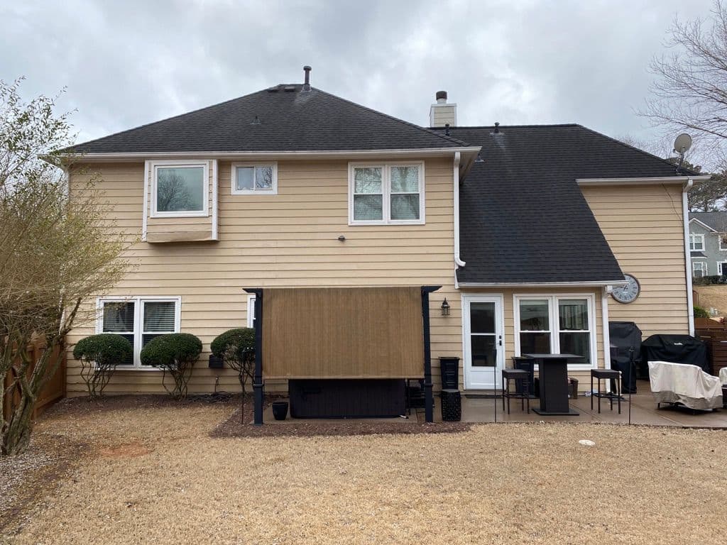 Two-story beige house with black roof, patio, chairs, grill, and a covered outdoor seating area. Overcast sky and bare trees suggest winter.
