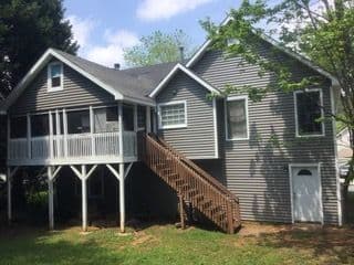 Two-story gray house with white trim, wooden stairs, and a screened porch. Surrounded by trees, it has a calm, suburban feel under a bright sky.