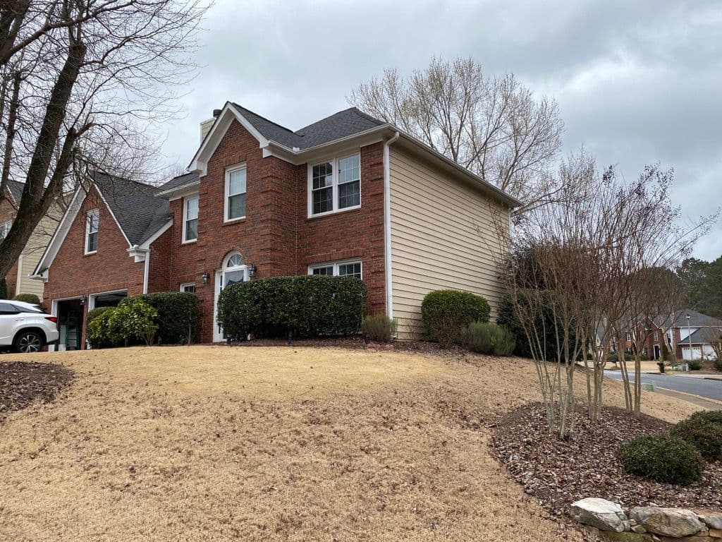 Brick suburban house on a hill with a manicured lawn and shrubs. Overcast sky casts a muted tone over the neighborhood.