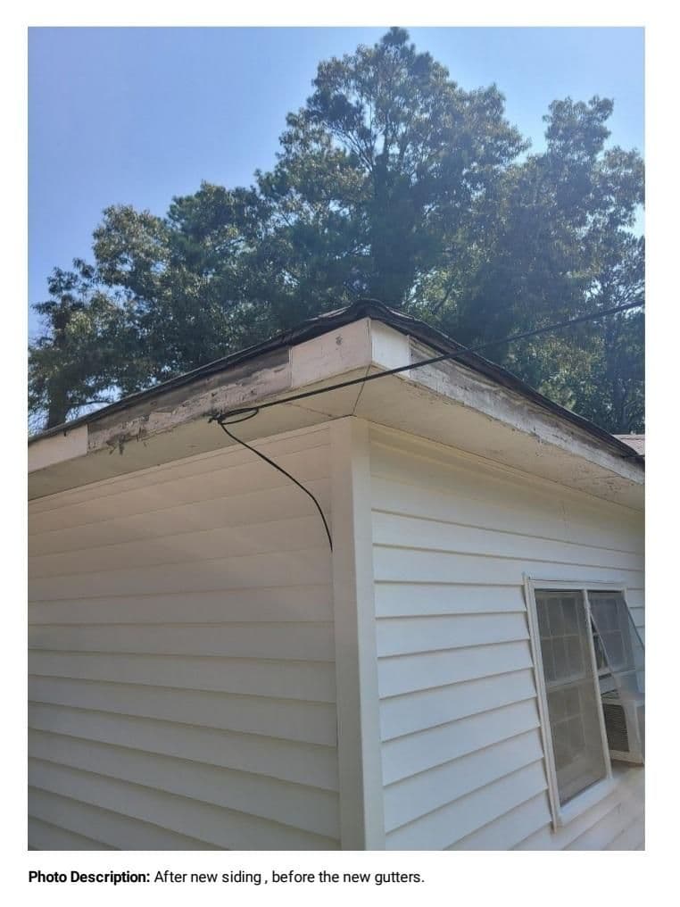 White house corner with new siding, showing missing gutters and exposed wires. Trees in the background under a clear blue sky.