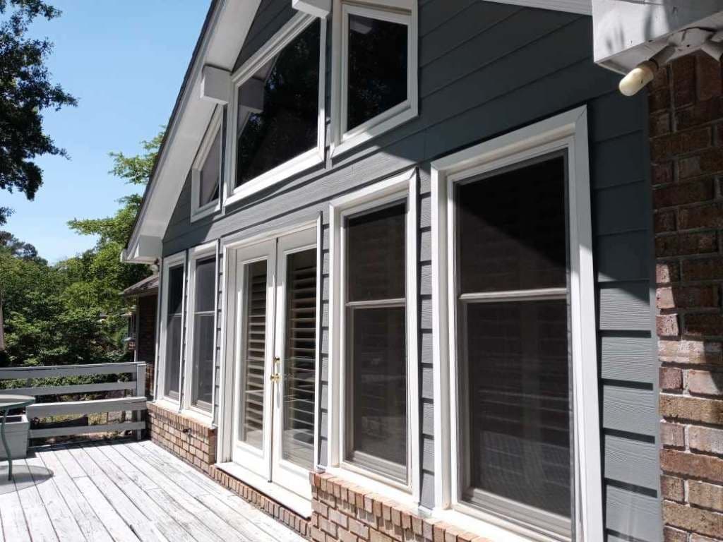A modern house exterior with large triangular windows, French doors, and gray siding. Sunlit wooden deck in front, surrounded by lush green trees.