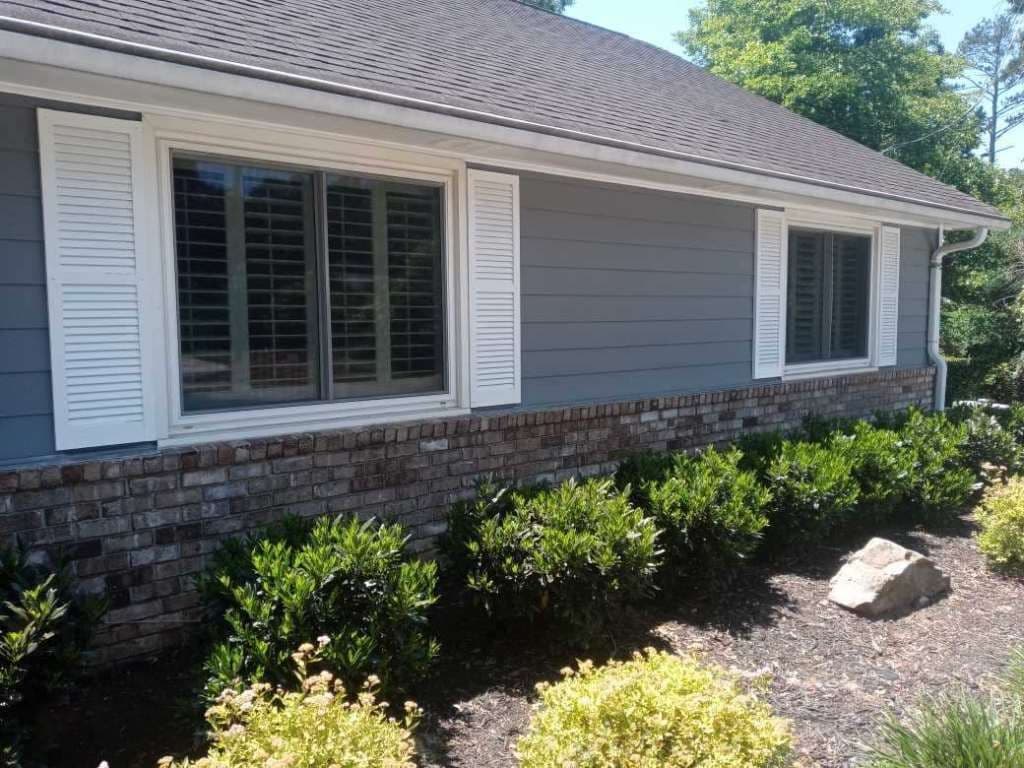 Side view of a house with light blue siding and white shutters, featuring two large windows. The lower section is red brick. Bushes and shrubs grow in front, under a clear blue sky.