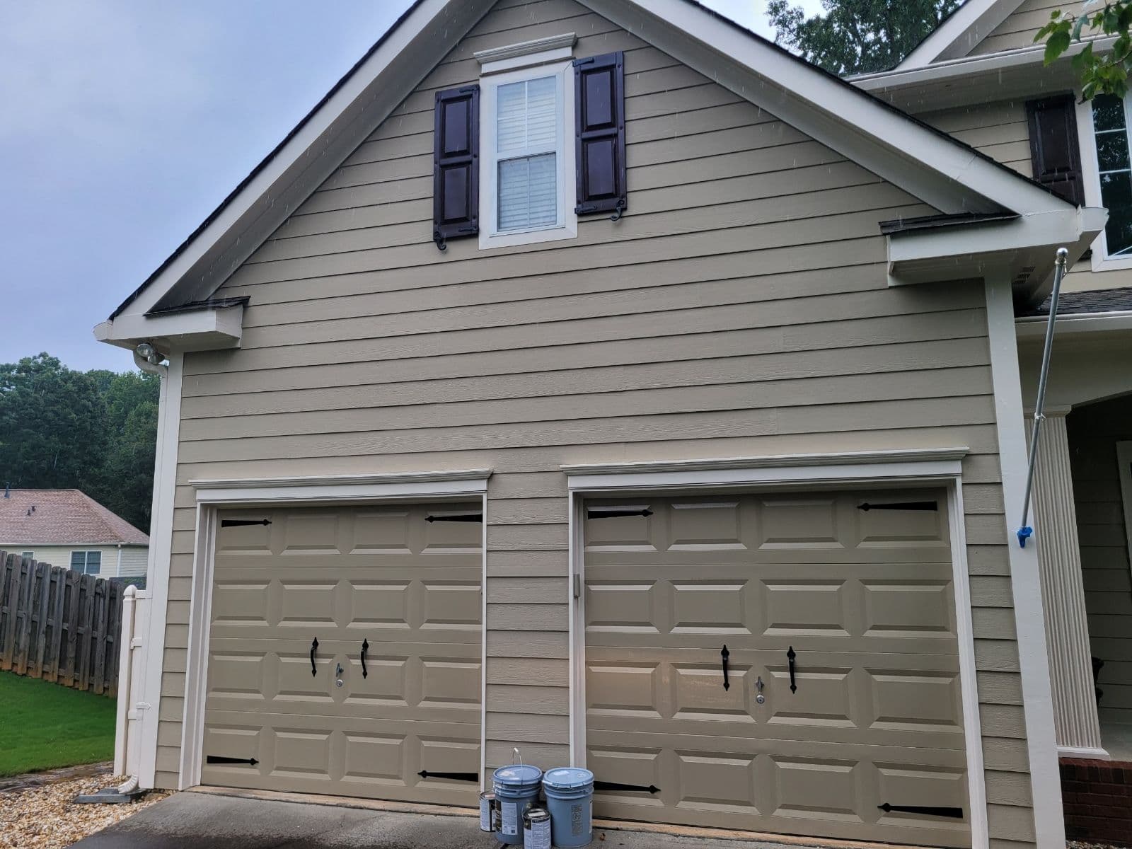 Tan, double garage with two closed doors and a small attic window. Below the window are two dark shutters. Blue paint cans sit on the driveway. Calm, overcast day.