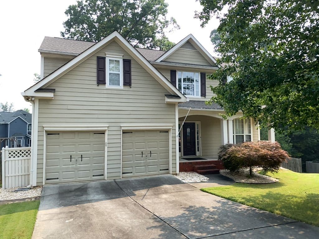 Suburban house with beige siding, dark shutters, and a two-car garage. Front porch has steps and is flanked by a tree and small landscaped garden.