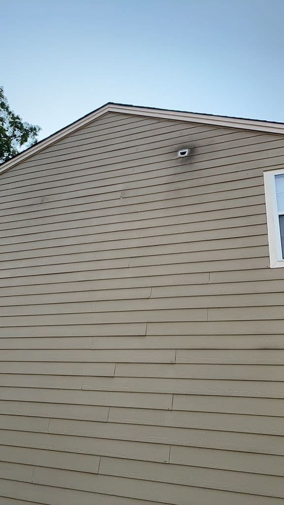 Side view of a tan house with horizontal siding, featuring a small security camera and a black smudge. Clear sky above, with a hint of green foliage.