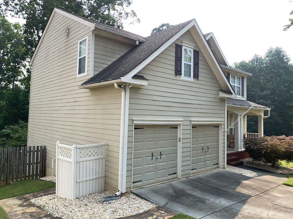 Two-story beige house with white trim, featuring a double garage, porch, and lush green surroundings. The setting is calm with a clear sky.