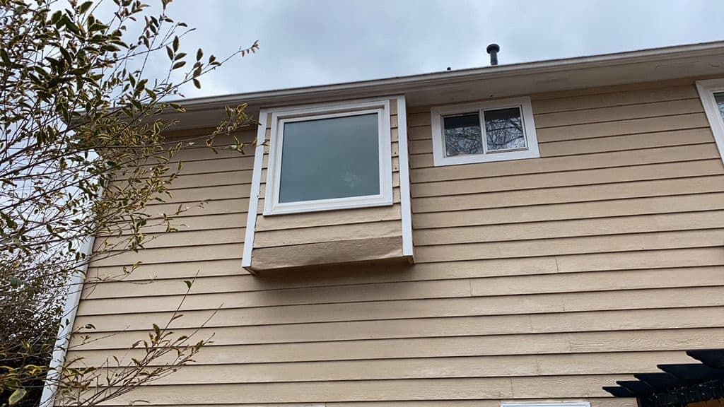 Tan siding house with large rectangular window protrusion on upper floor, adjacent smaller window. Overcast sky and branches visible.