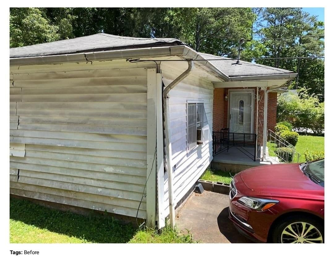 A small, worn house with peeling white siding and a sagging roof stands in sunlight. A red car is parked to the side. The setting is tranquil yet neglected.