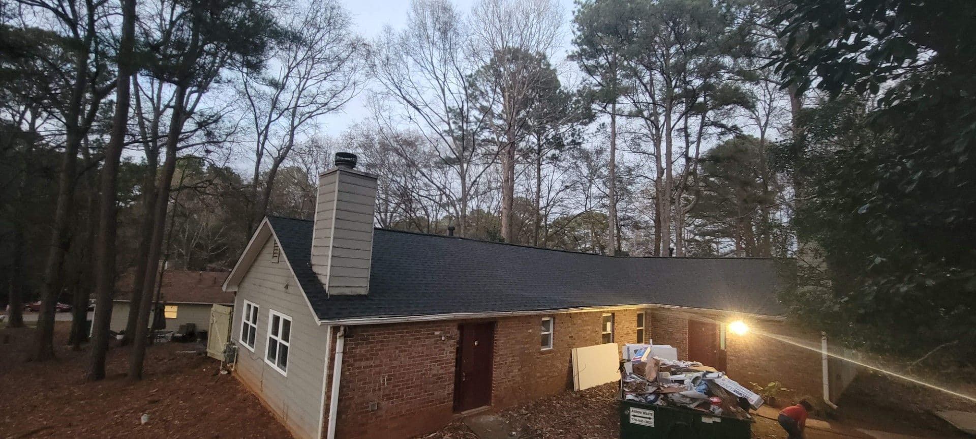 A brick house with a black roof is surrounded by tall trees under a cloudy sky. A light by the door illuminates a dumpster filled with construction debris.