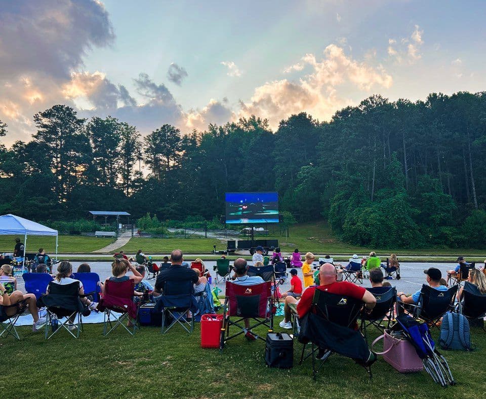 Outdoor movie night with people in folding chairs watching a screen at dusk. The background features lush trees and a cloudy sky, creating a relaxed atmosphere.