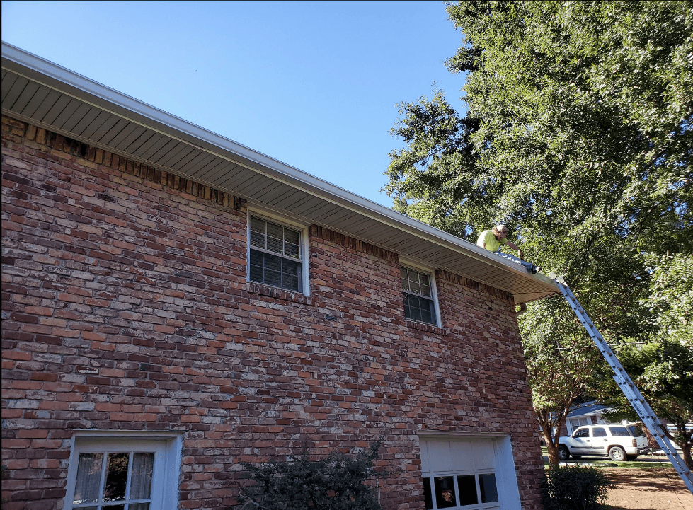 A person is on a ladder cleaning the gutters of a two-story brick house. The scene is sunny, and the home is surrounded by lush green trees.