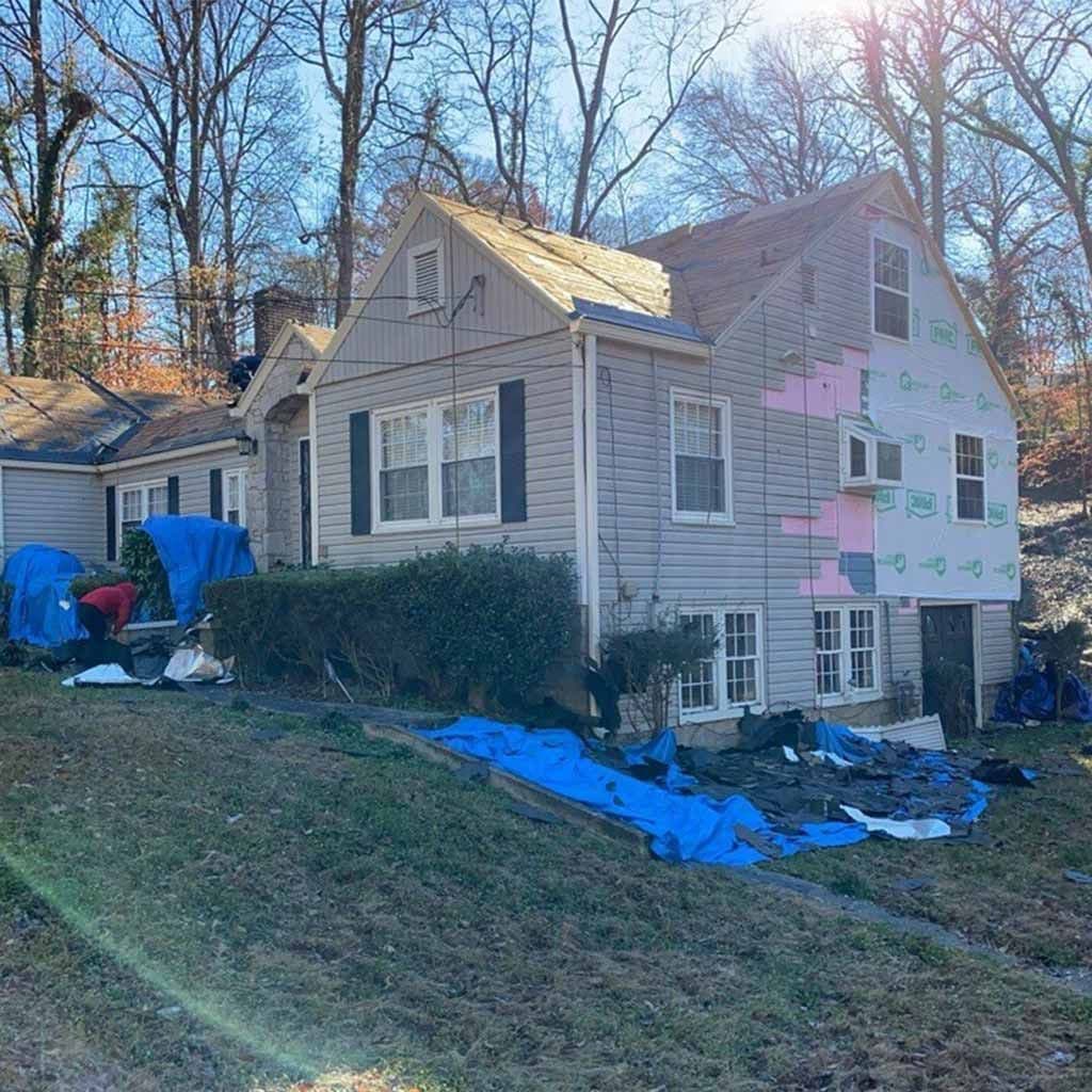 A gray house undergoing repairs, with several blue tarps covering parts of the roof and siding. A person works near bushes, and tall trees are in the background.