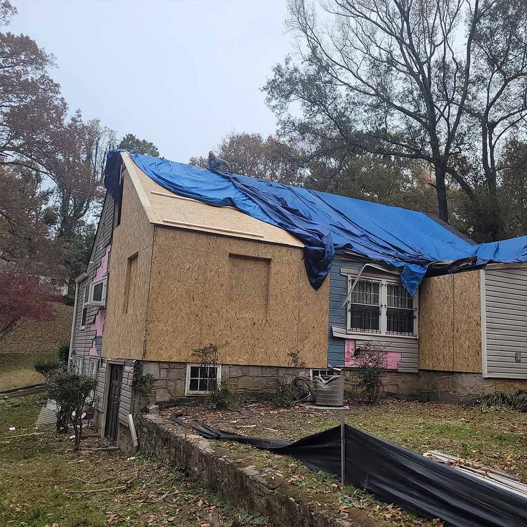 House under renovation with plywood walls and a blue tarp covering the roof. Surrounded by trees and a yard, conveying an ongoing construction feel.