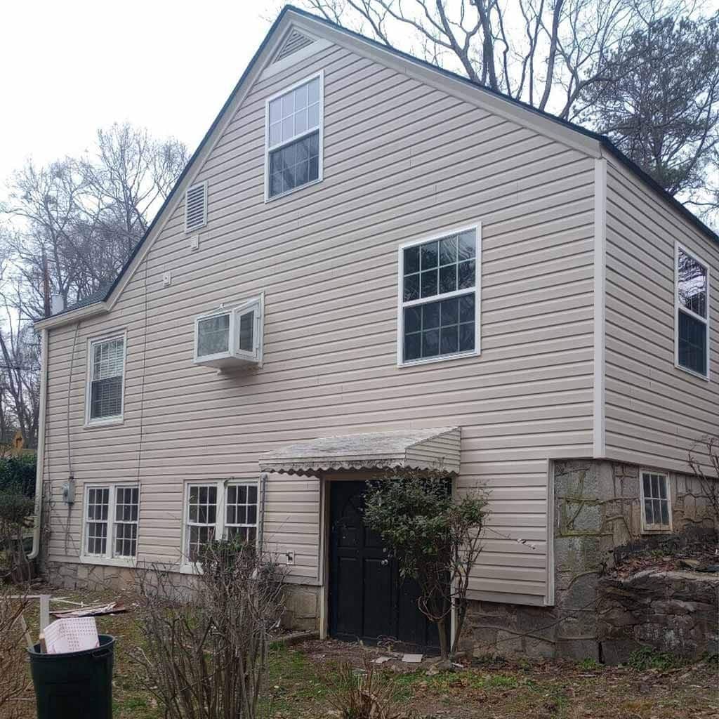 A beige-sided house with multiple windows, a black door, and a small awning. Leafless trees and a trash can are in the front yard on a cloudy day.