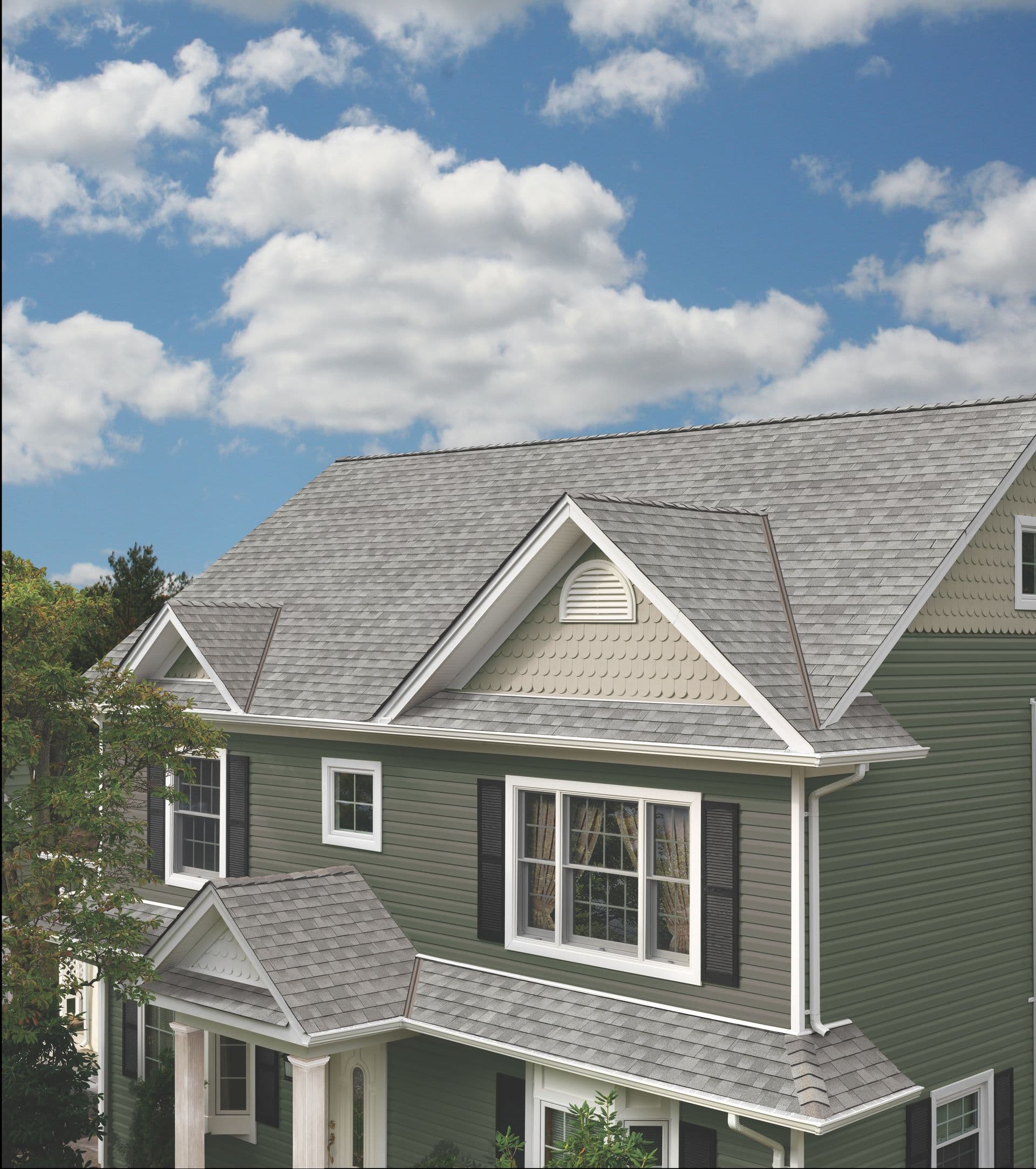 A two-story house with green siding and grey roof sits under a blue sky with fluffy clouds. Large windows and white trim convey a welcoming feel.