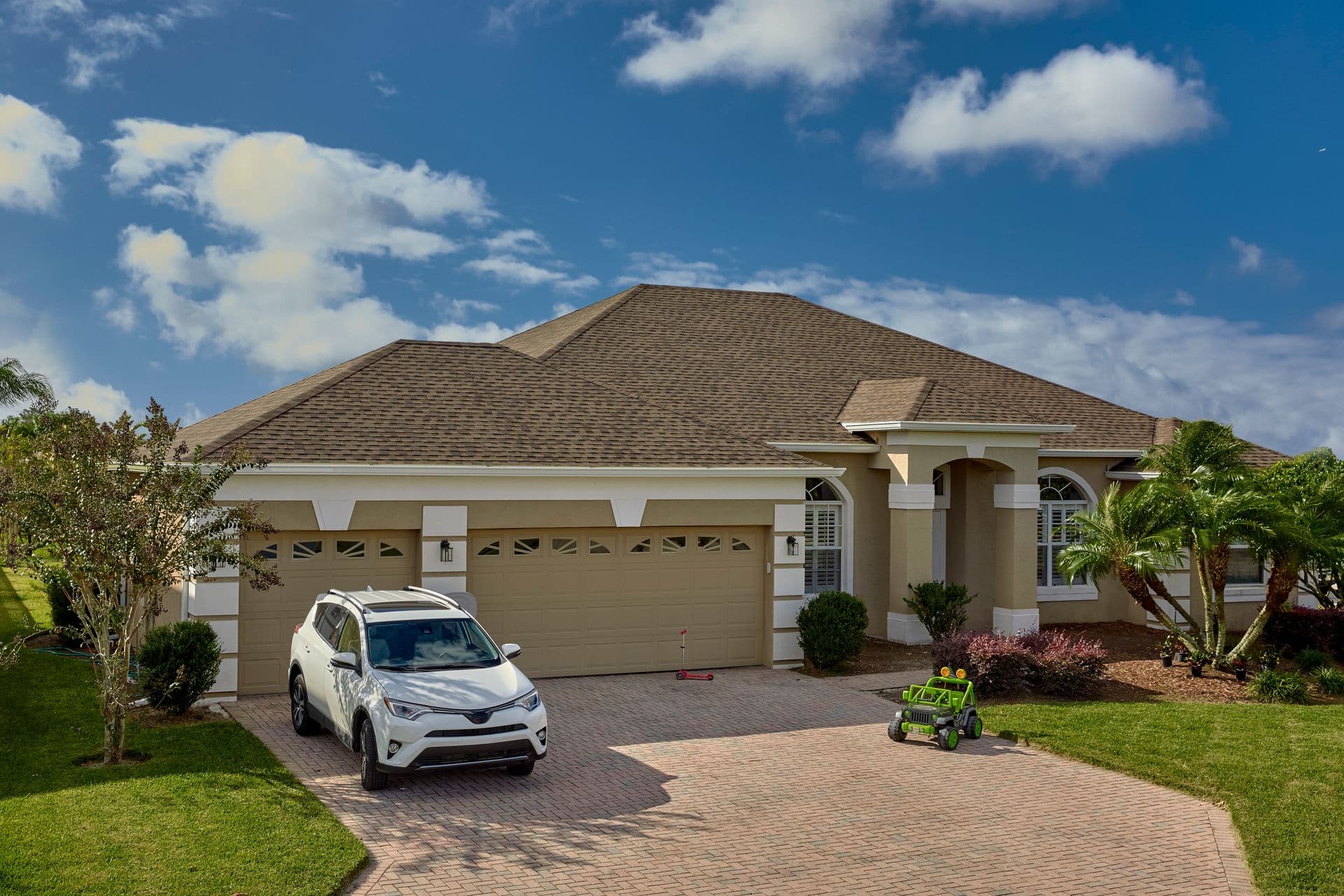 A modern suburban home with a beige exterior, wide garage, and a well-kept garden. A white SUV is parked in the driveway under a blue sky.