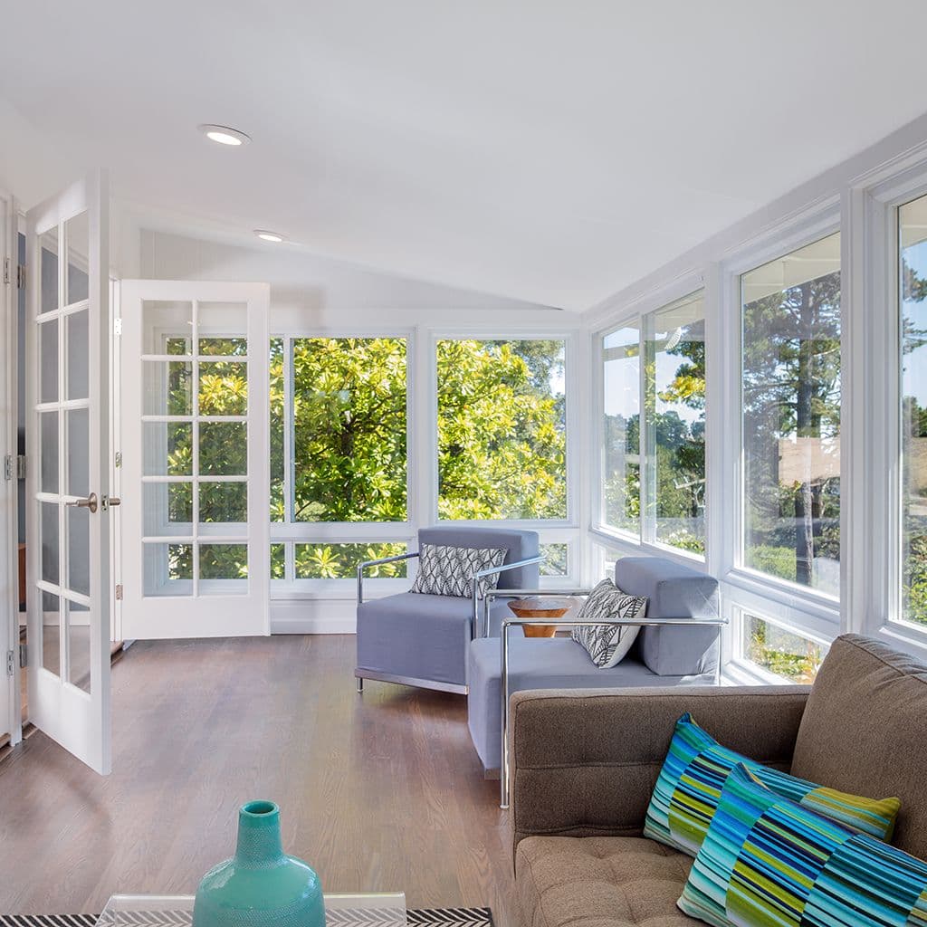 A bright sunroom with large windows and green views. Modern gray chairs, a beige sofa with colorful pillows, and light wood flooring create a serene vibe.