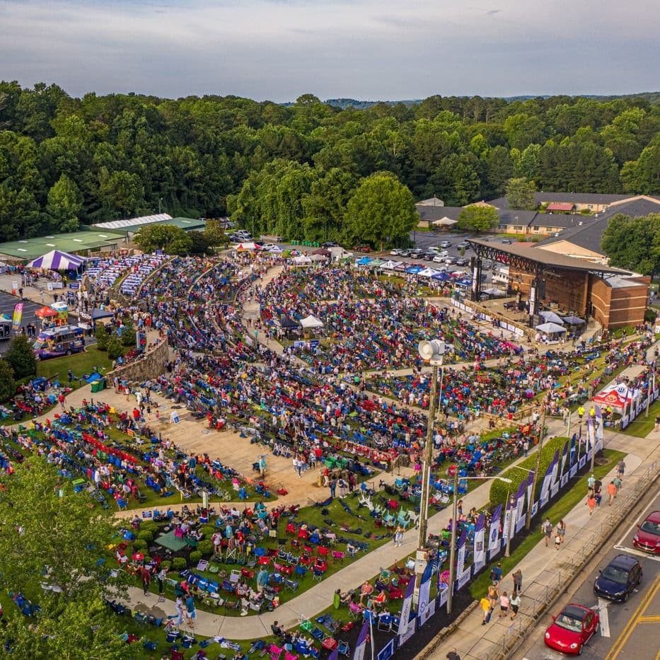 Aerial view of a vibrant outdoor concert at an amphitheater. Hundreds of people in colorful chairs fill the venue, surrounded by lush green trees.