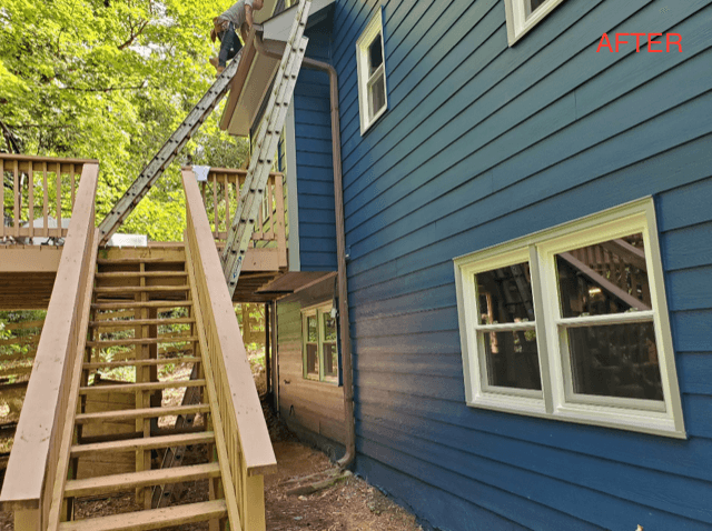 Blue house exterior with three white windows; a person on a ladder near the gutter. Wooden stairs lead to a deck. Verdant trees in the background.